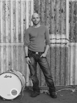 Mann steht vor Wellblechwand, neben ihm eine Trommel.