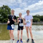 Drei junge Läuferinnen mit Medaillen posieren vor einem Teich mit Startnummern.