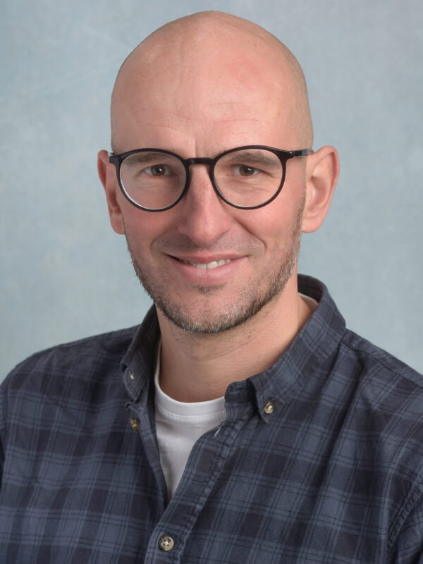 Ein lächelnder Mann mit Brille und kariertem Hemd vor grauem Hintergrund.