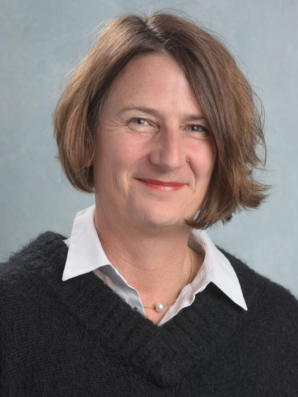 Eine lächelnde Person mit braunem Haar trägt eine weiße Bluse und einen schwarzen Pullover.