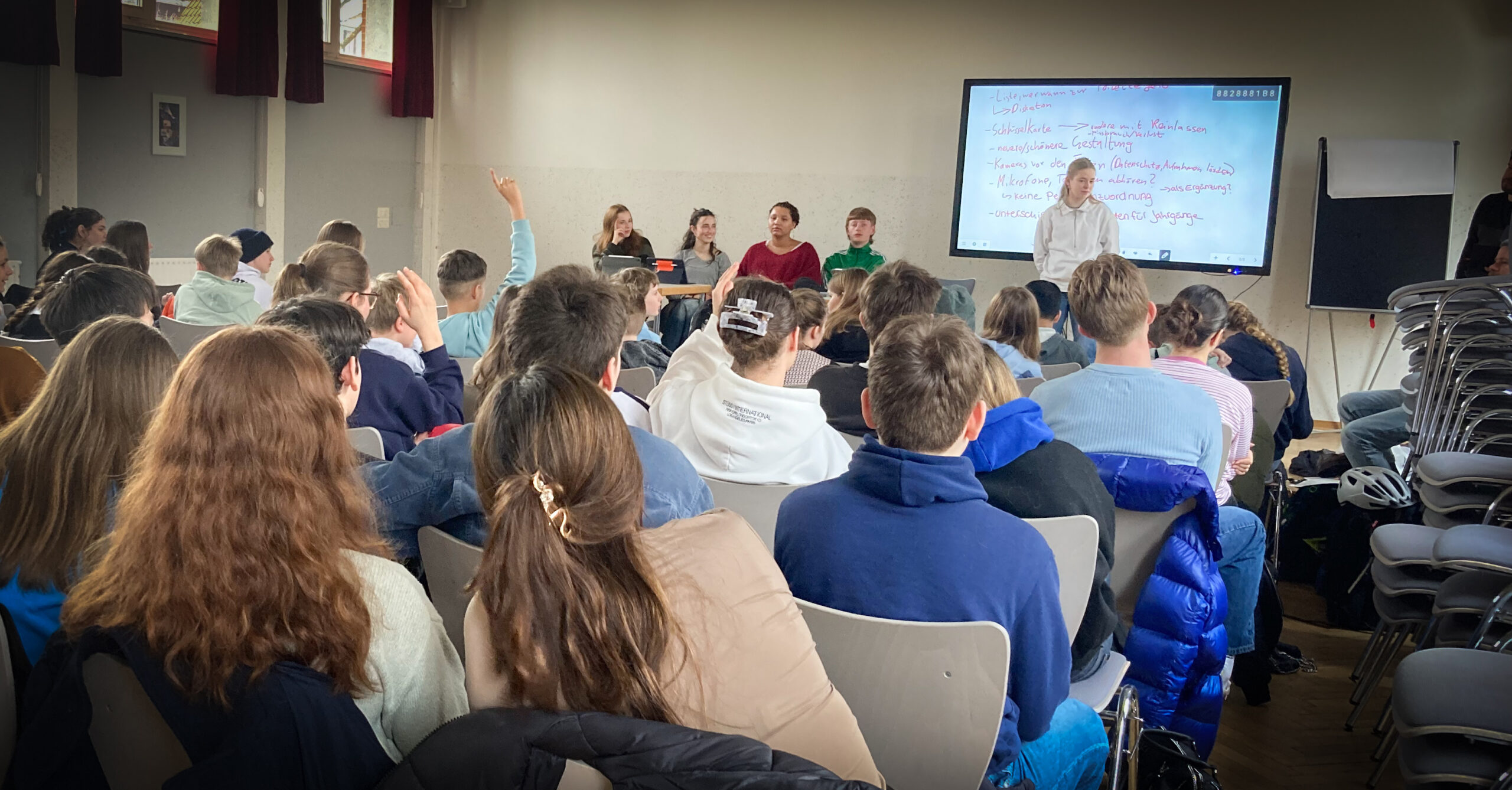 Schüler sitzen bei einem Vortrag oder einer Diskussion in einem Klassenraum, einige melden sich.