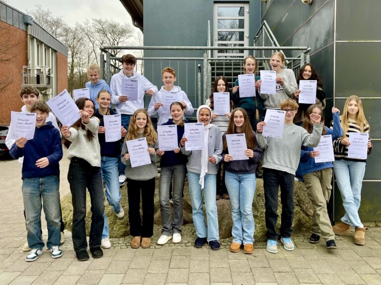 Gruppe Jugendlicher hält Urkunden vor einem Schulgebäude und lächelt in die Kamera.