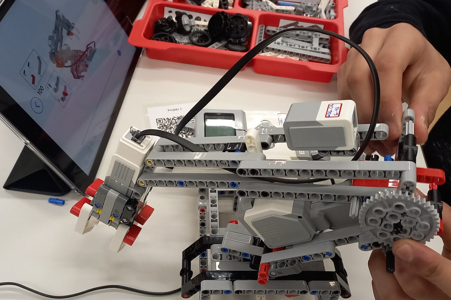 Person baut mit Lego-Robotikset, Tablet zeigt Bauanleitung, bunte Legosteine im Hintergrund.