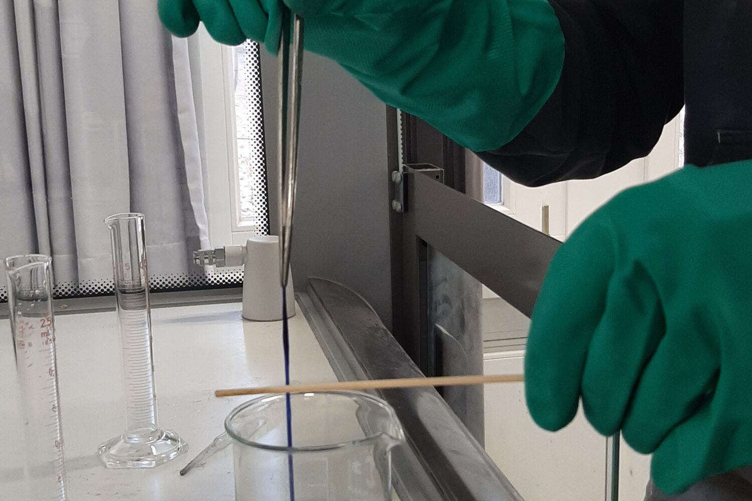 Person mit grünen Handschuhen rührt blaue Flüssigkeit in einem Becherglas um.