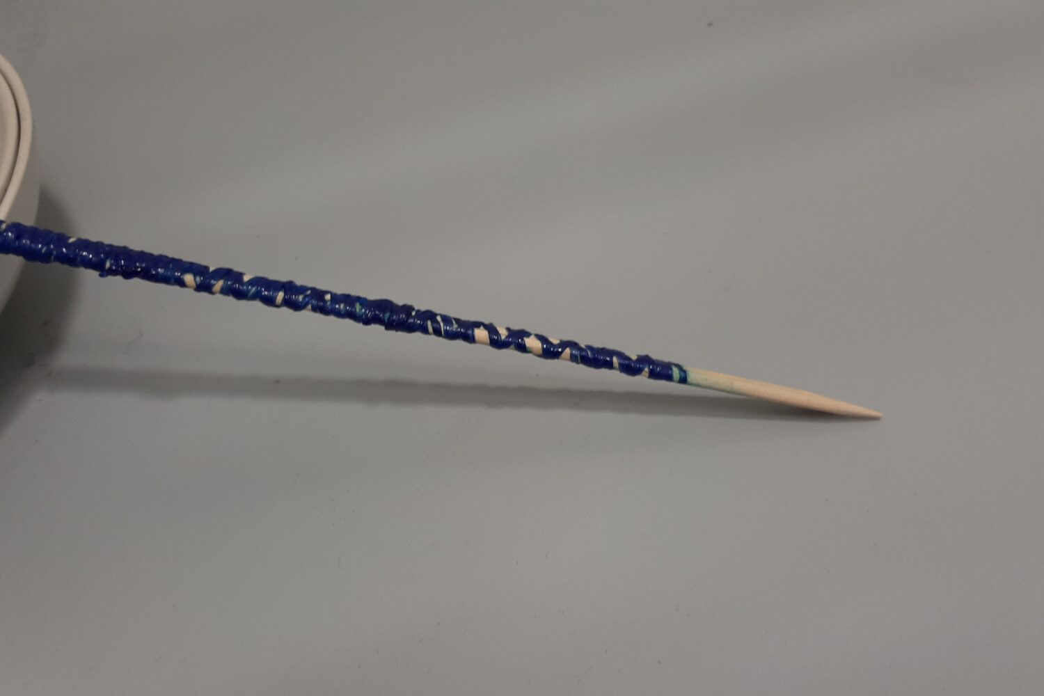 Ein Zahnstocher mit blauer Farbe bestrichen, liegt auf einem grauen Tisch neben einer Schale.