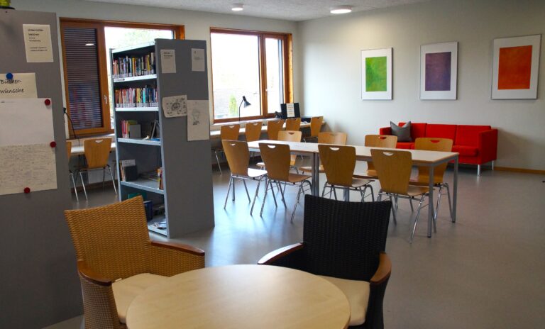 Moderner Raum mit Tischen, Stühlen, rotem Sofa und Bücherregal. Helle Fenster.