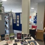 Ein Tisch mit verschiedenen Büchern in einer Bibliothek, umgeben von Regalen und Stühlen.