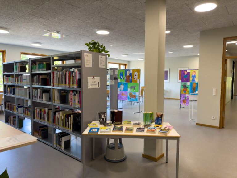 Helle Bibliothek mit Regalen, Tisch voller Bücher und bunten Kinderkunstwerken im Hintergrund.
