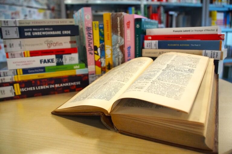 Aufgeschlagenes Buch vor bunt gestapelten Büchern auf einem Tisch in einer Bibliothek.