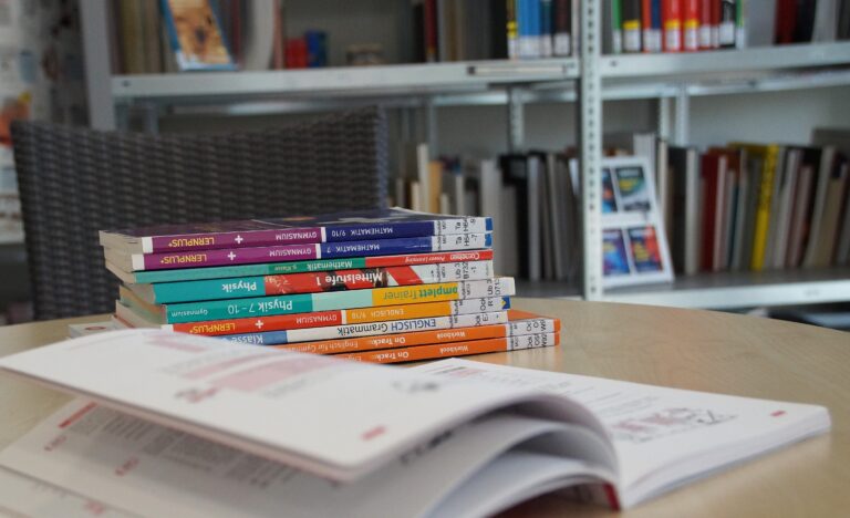Stapel bunter Schulbücher auf Tisch, offenes Buch im Vordergrund, Bücherregal im Hintergrund.
