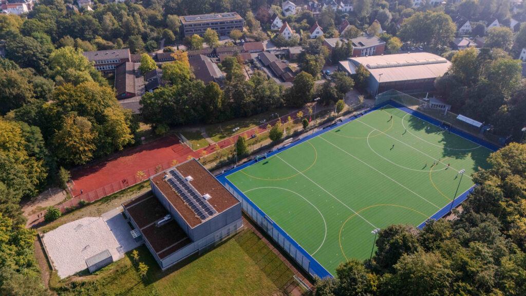 Luftaufnahme eines Sportplatzes und Umgebung, mit Bäumen und Gebäuden im Hintergrund.