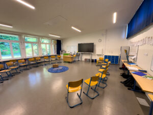 Klassenzimmer mit Stuhlkreis, Pult, Whiteboard und Blick in den Garten durch große Fenster.