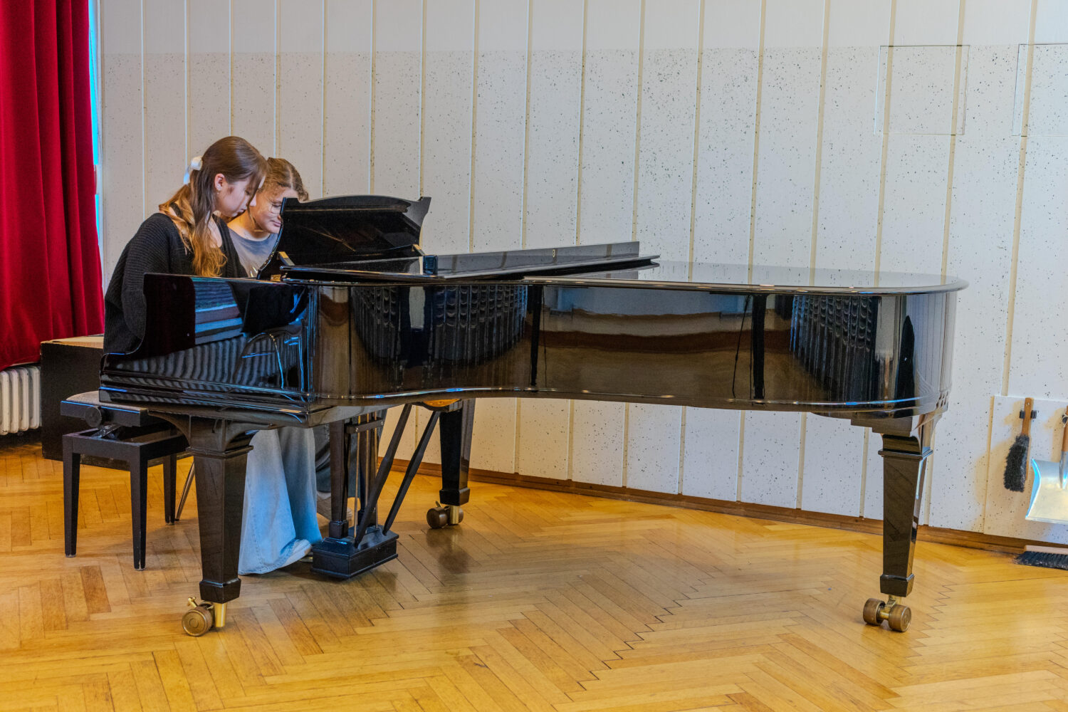 Zwei Personen spielen gemeinsam am schwarzen Flügel in einem Raum mit Holzboden.