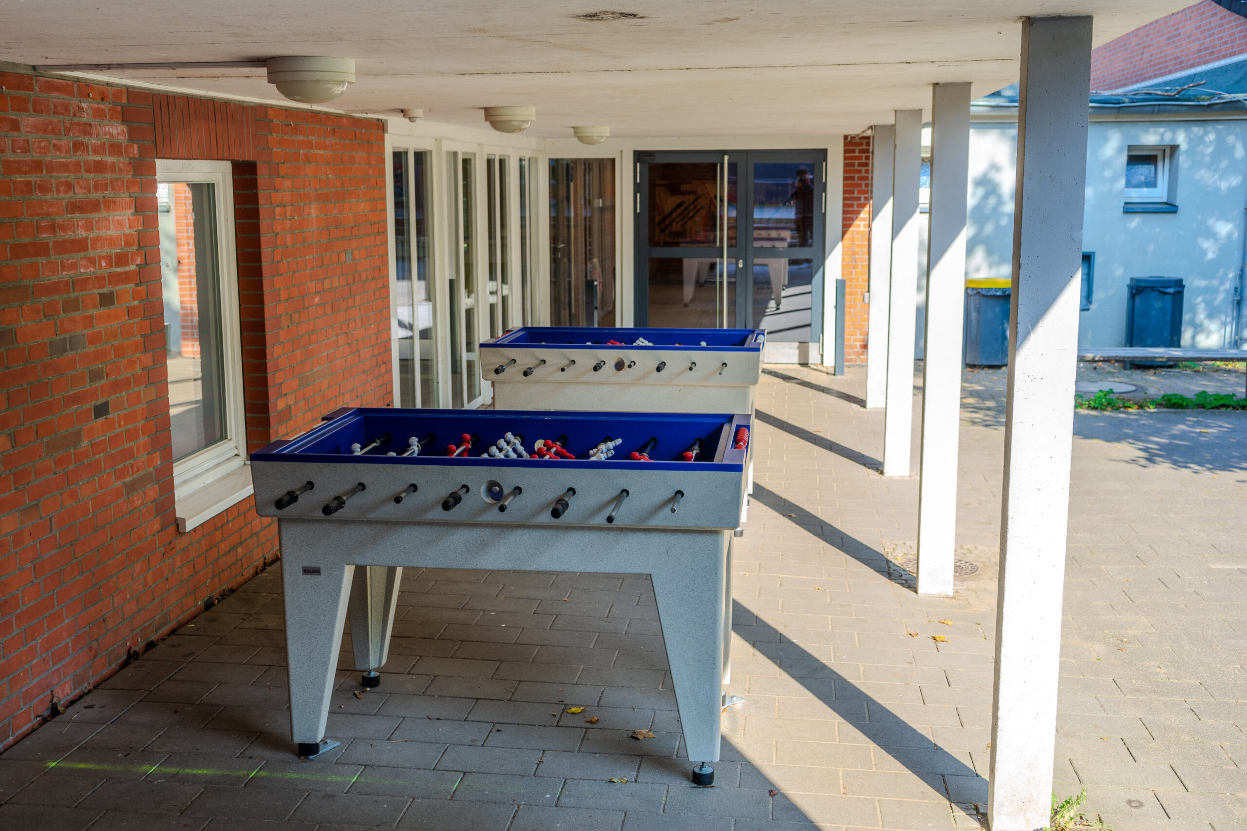 Zwei Tischfußballtische stehen auf einer überdachten Terrasse vor einem Gebäude.