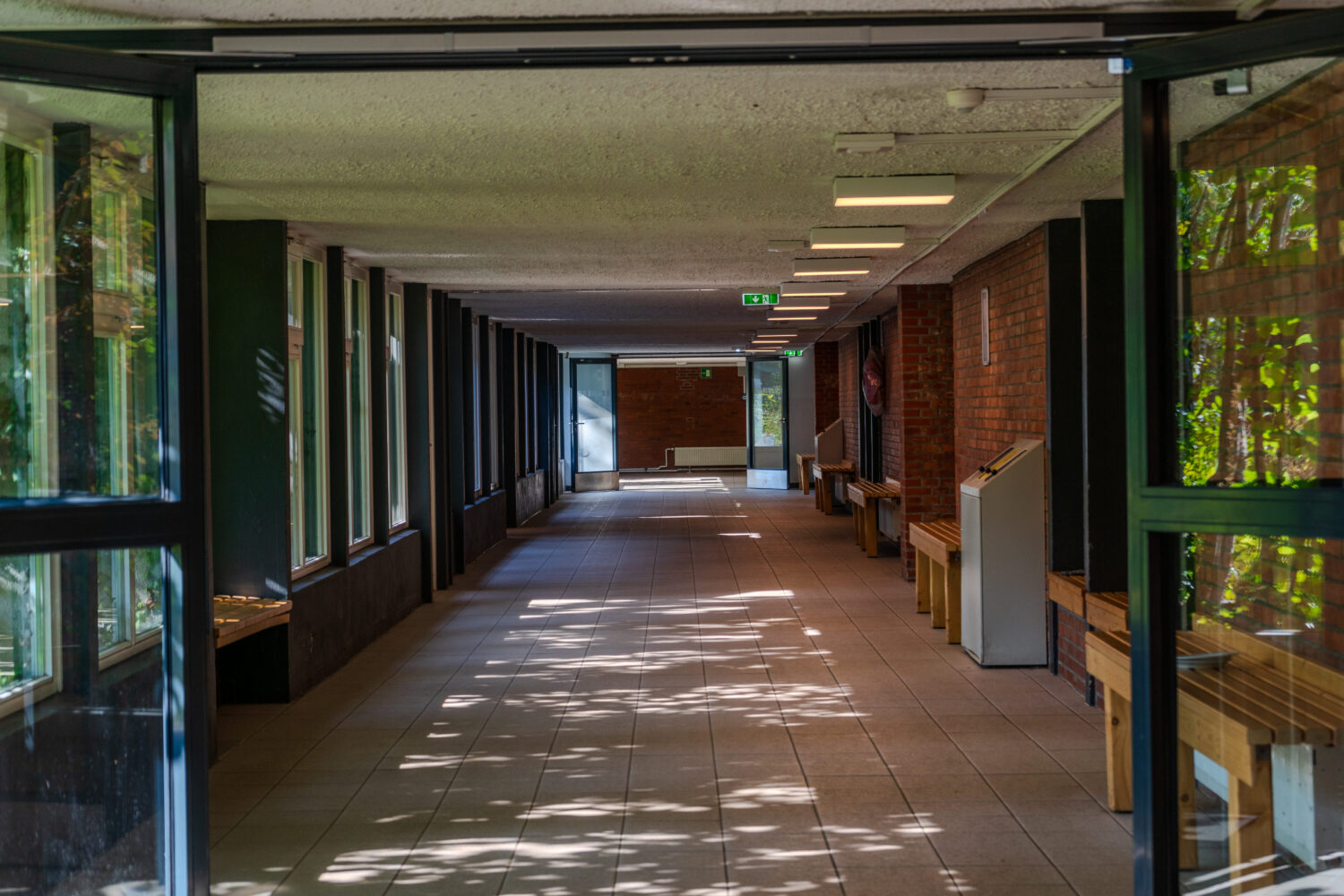 Langer Flur mit Fenstern links, Bänken rechts und Schattenmustern am Boden.