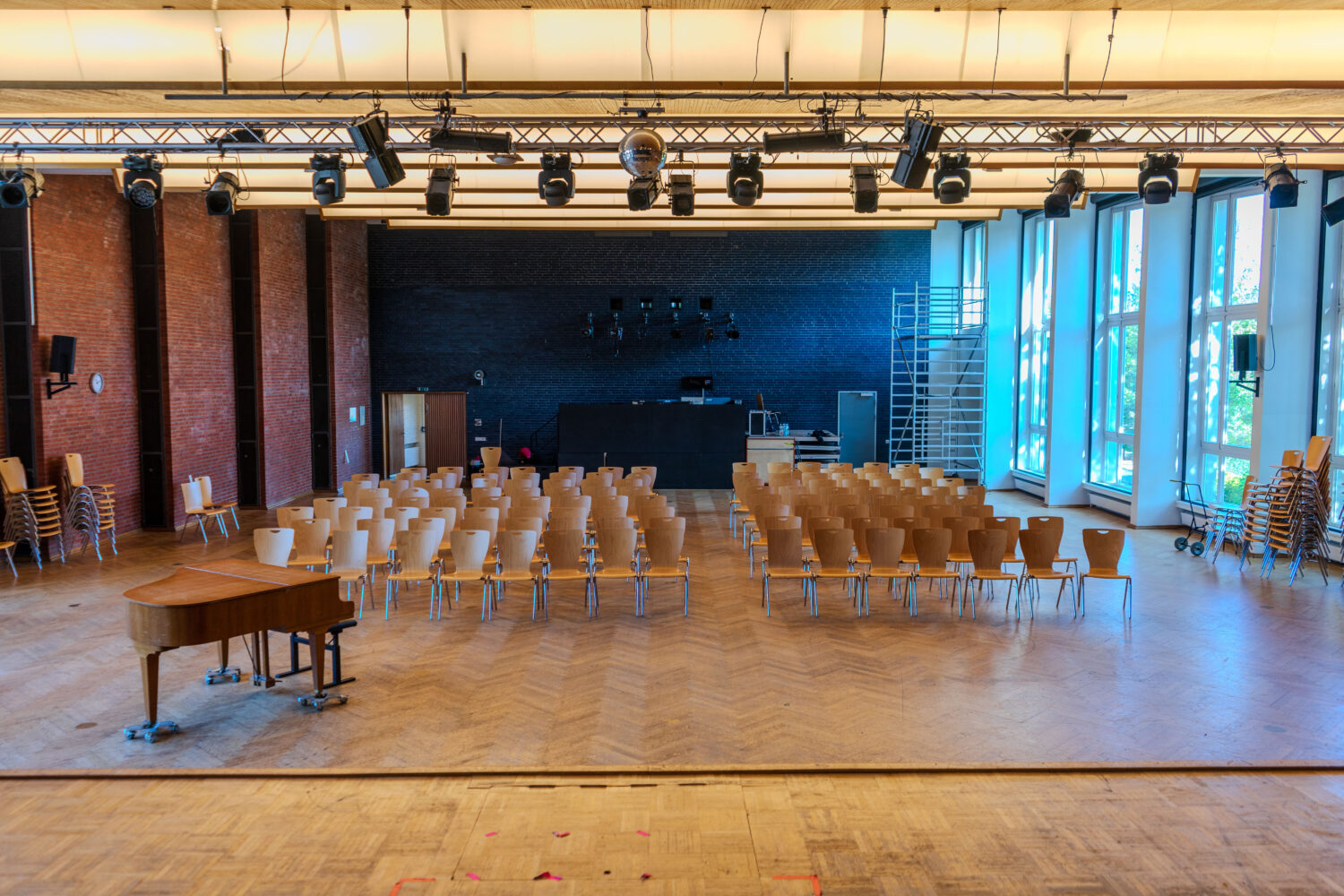 Großer Saal mit gestapelten Stühlen, Flügel und heller Beleuchtung.