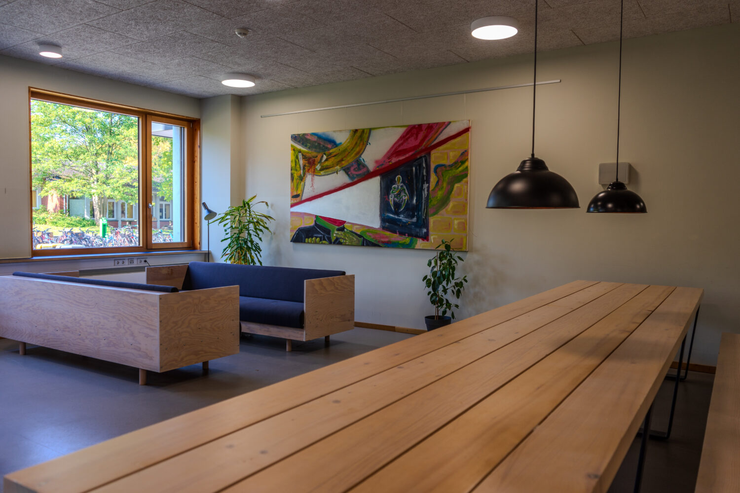 Moderner Raum mit Holzbank, Gemälde, Pflanzen und großer Fensteransicht auf grüne Bäume.