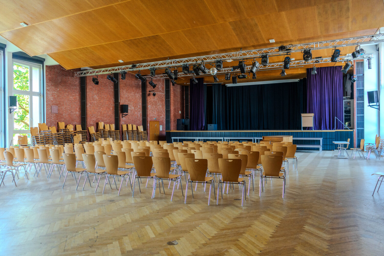 Großer Saal mit Holzboden, Bühne und gestapelten Stühlen.
