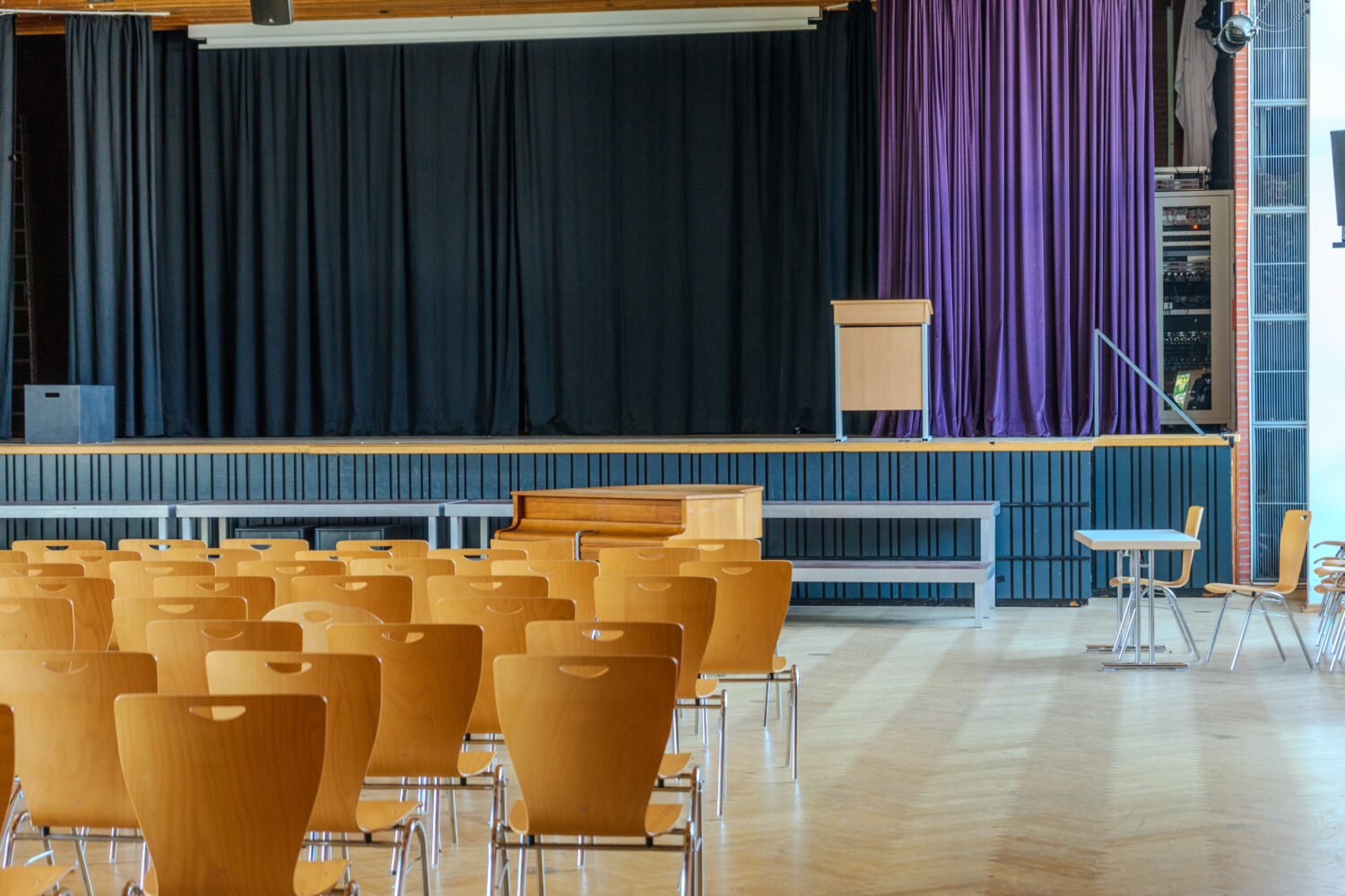 Leerer Saal mit Reihen von Stühlen vor einer Bühne mit schwarzem Vorhang und Rednerpult.