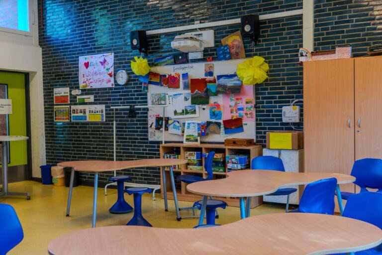 Heller Klassenraum mit runden Tischen, blauen Stühlen und bunter Wanddekoration.