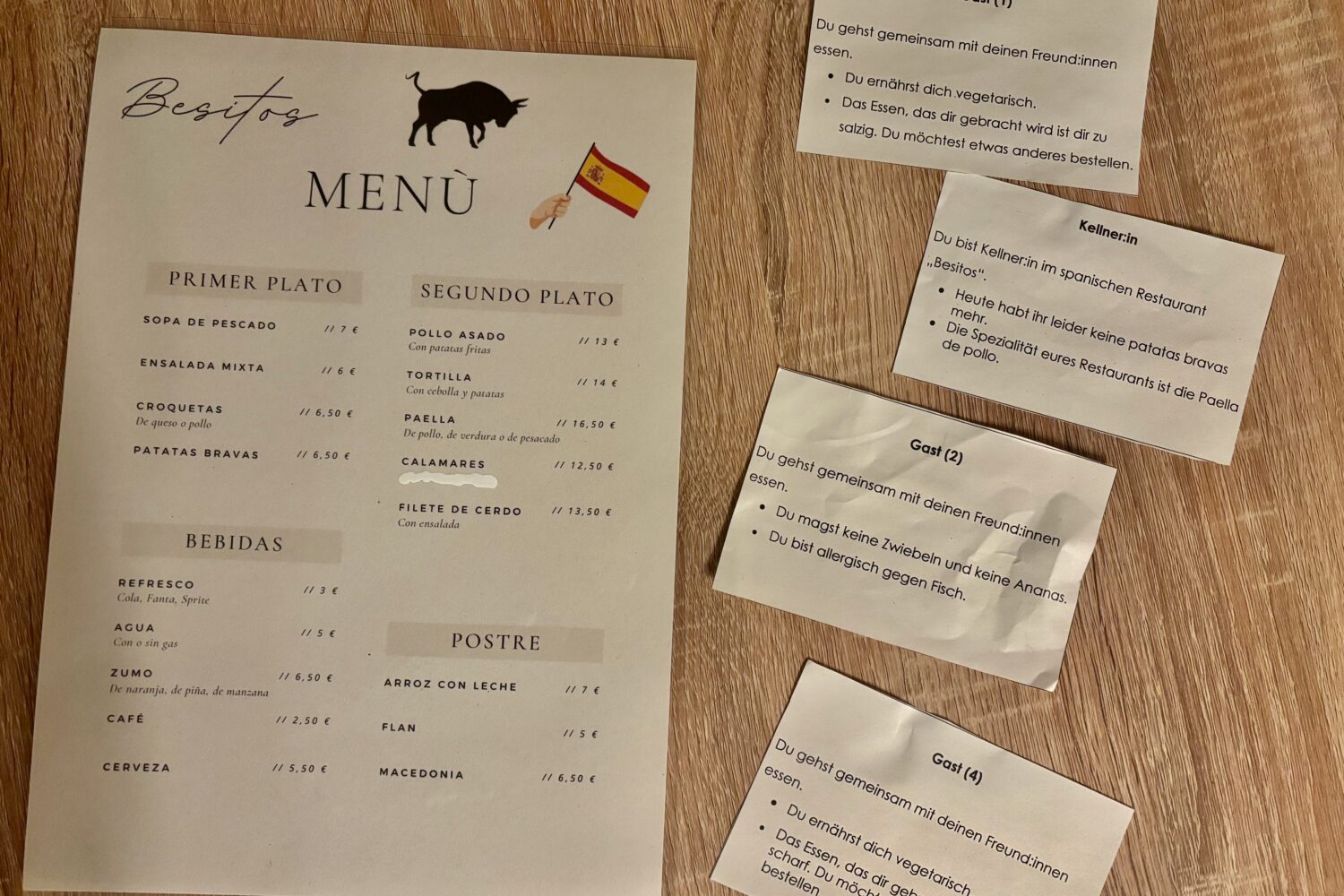 Spanisches Menü und Notizen mit Gäste- und Kellner:in-Rollen auf Holztisch.