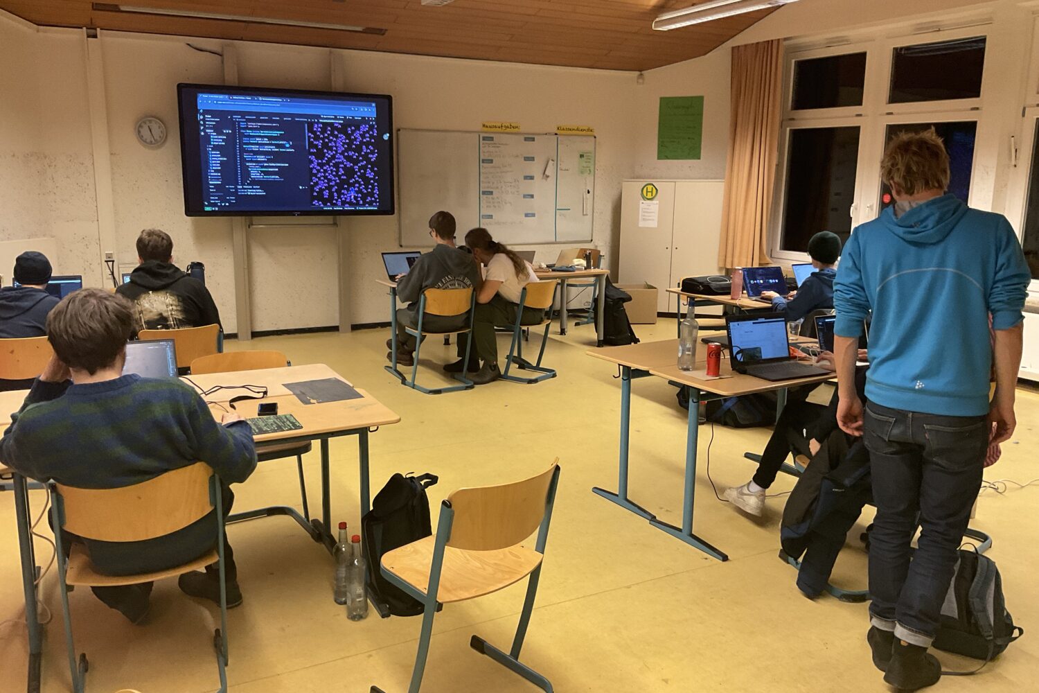 Schüler arbeiten in einem Klassenzimmer an Laptops, großer Bildschirm zeigt Code an.