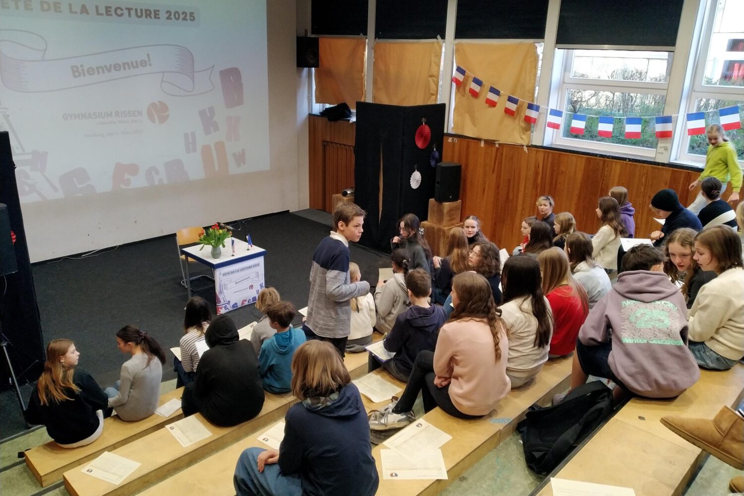 Schüler in Auditorium, warten auf Veranstaltung. Fête de la Lecture 2025 auf Leinwand.
