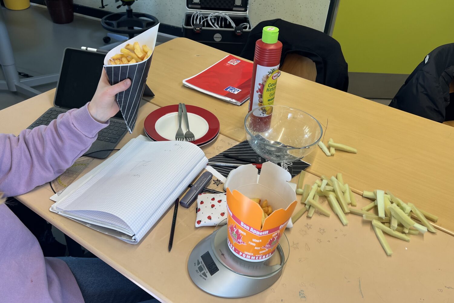 Person hält Pommes, Waage misst Gewicht, Notizbuch, Pommes auf Tisch, Ketchup-Flasche daneben.