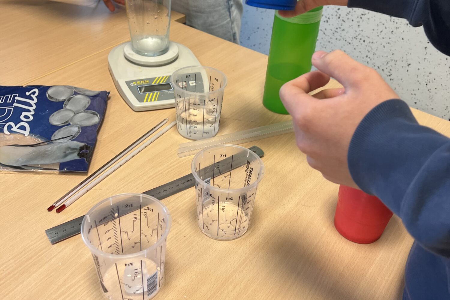 Personen wiegen Flüssigkeit mit Messbechern, einem Becher und einer Waage auf einem Tisch ab.