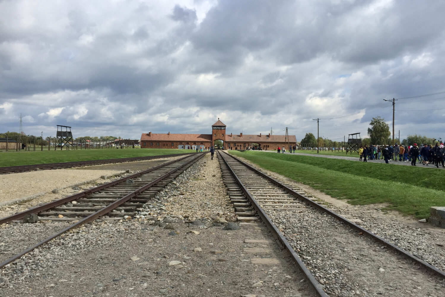 Eisenbahnschienen führen zu einem Tor mit Wachturm, links und rechts Menschen auf einer Wiese.