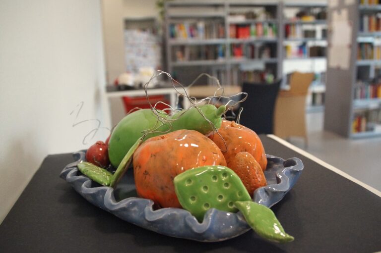 Keramikfrüchte auf blauem Teller mit Drahtdeko, im Hintergrund unscharfe Bibliothek.