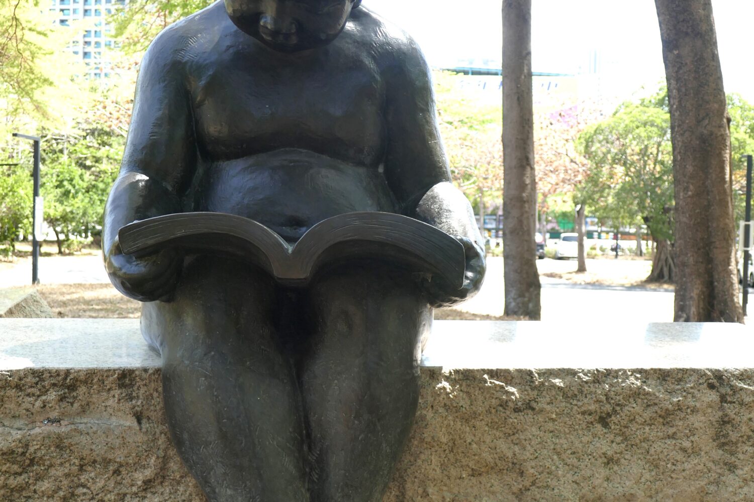 Bronzestatue eines lesenden Kindes auf einer Steinbank im Park.