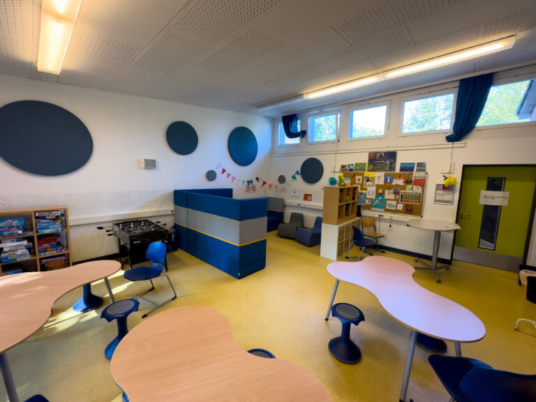 Moderner Klassenraum mit runden Tischen, bunten Stühlen und Pinnwand an der Wand.