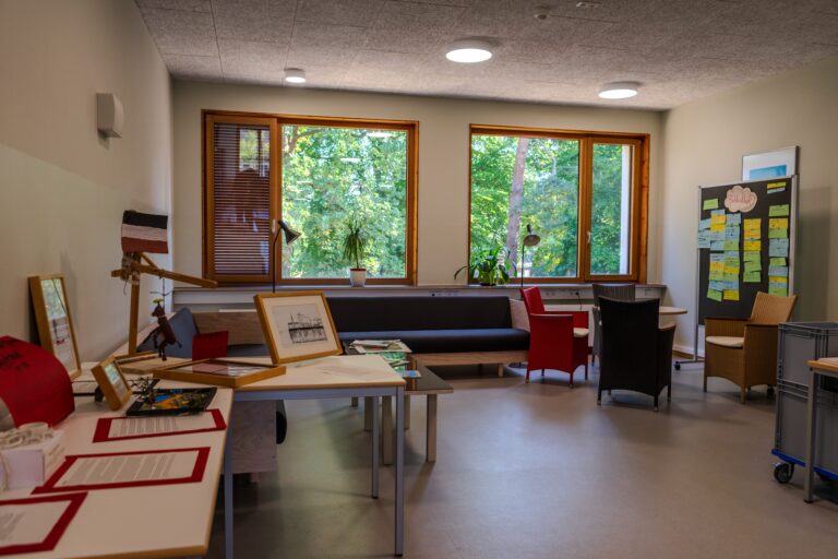Ein helles Zimmer mit Tischen, Stühlen und viel Grün draußen vor den Fenstern.