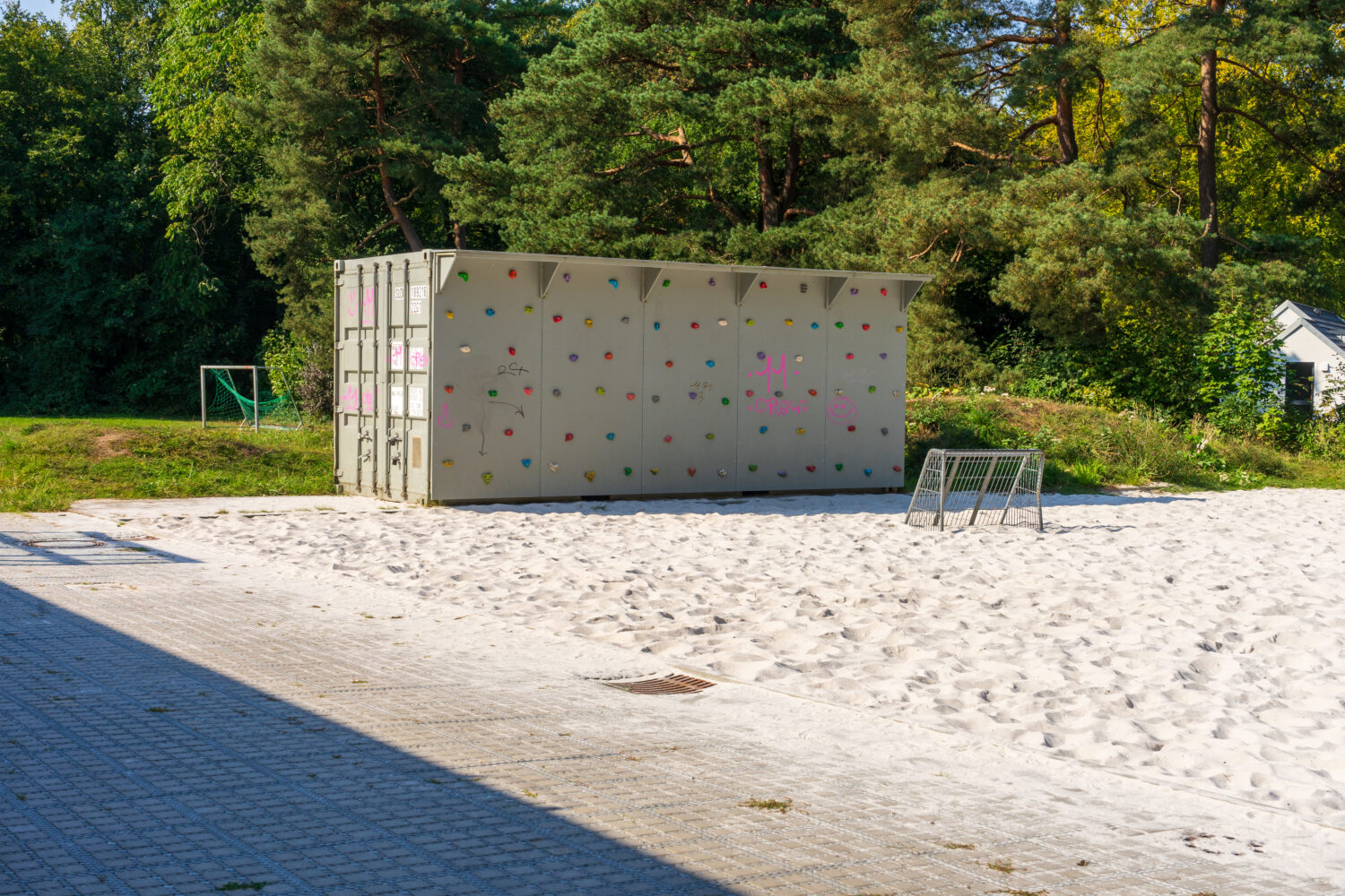 Kletterwand auf Container im Sand vor einem kleinen Fußballtor, mit Bäumen im Hintergrund.