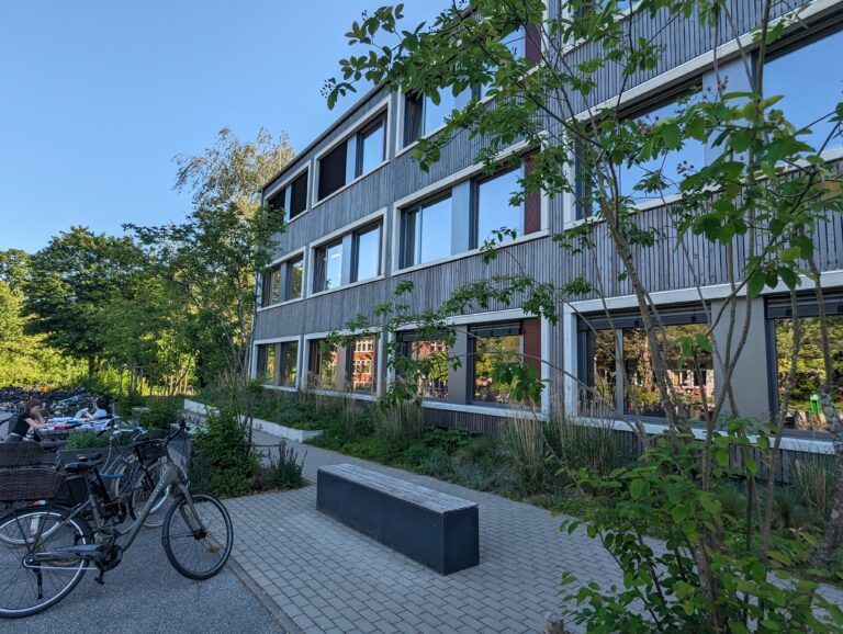 Modernes Gebäude mit großen Fenstern, umgeben von Bäumen und Fahrrädern bei sonnigem Wetter.