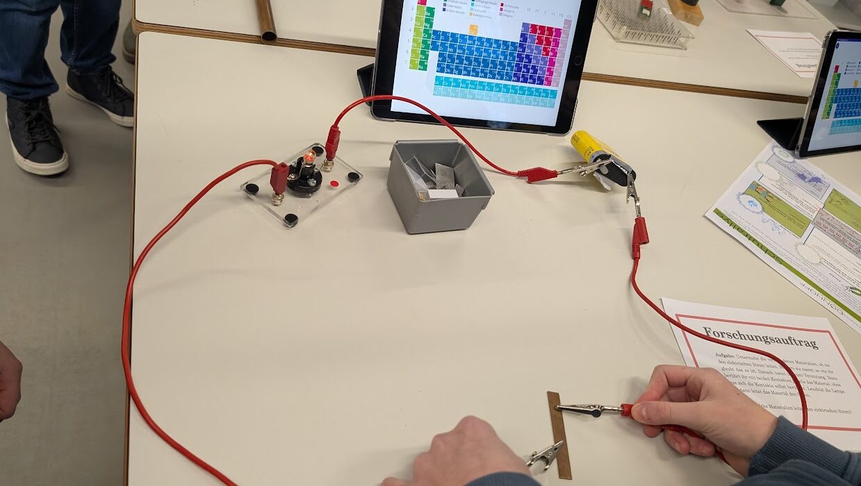 Person arbeitet mit Experimentieraufbau mit Kabeln, Krokodilklemmen und einem Tablet mit Periodensystem.