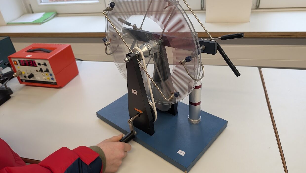 Person dreht an einer Wimshurst-Maschine auf einem Tisch; daneben ein rotes Messgerät.