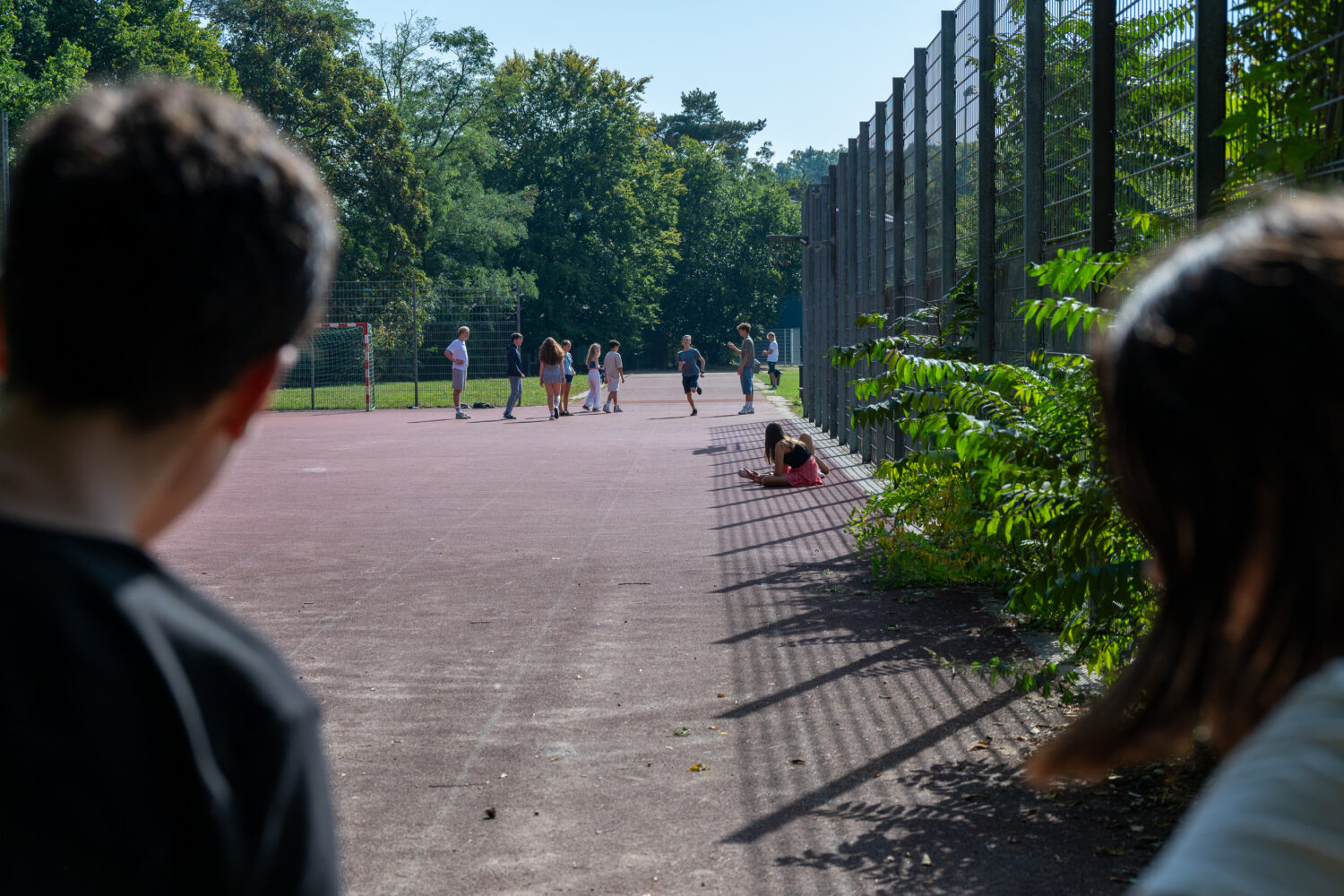Kinder spielen auf einem Sportplatz; ein Kind sitzt am Rand auf dem Boden.