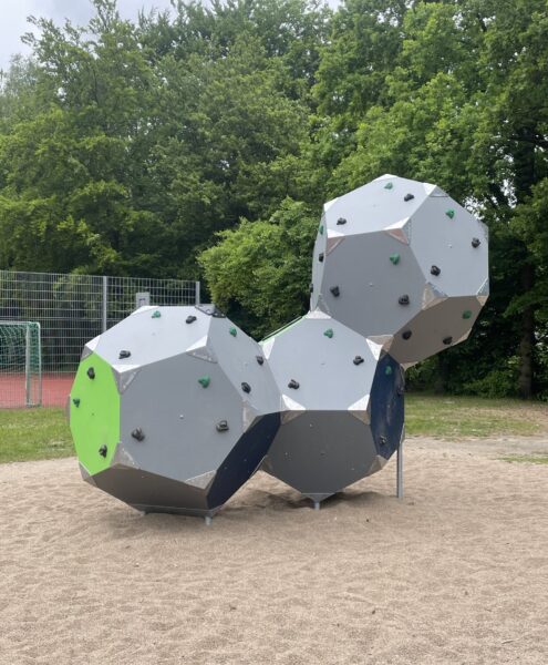 Drei graue Kletterelemente in geometrischer Form mit farbigen Griffen auf einem Spielplatz.