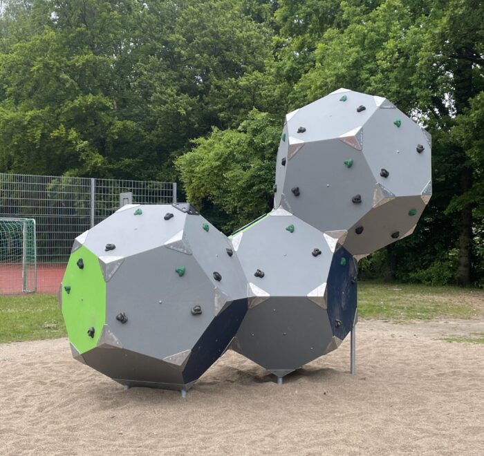 Drei graue Kletterelemente in geometrischer Form mit farbigen Griffen auf einem Spielplatz.