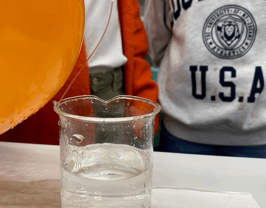 Oranger Ballon und Messbecher mit Wasser, daneben zwei Personen in Freizeitkleidung.