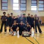 Jugendvolleyballteam jubelt in Sporthalle mit Anzeigetafel, zwei Erwachsene daneben.