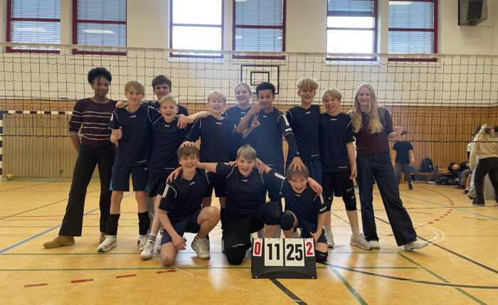 Jugendvolleyballteam jubelt in Sporthalle mit Anzeigetafel, zwei Erwachsene daneben.