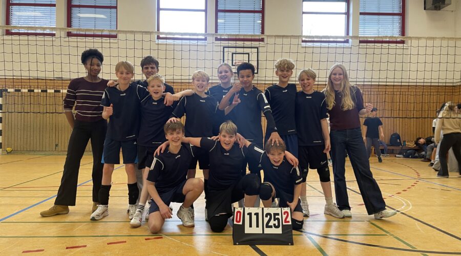 Jugendvolleyballteam jubelt in Sporthalle mit Anzeigetafel, zwei Erwachsene daneben.