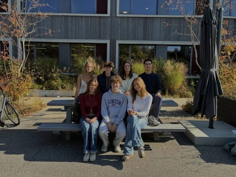 Eine Gruppe von sieben Personen sitzt lächelnd draußen an einem Holztisch.