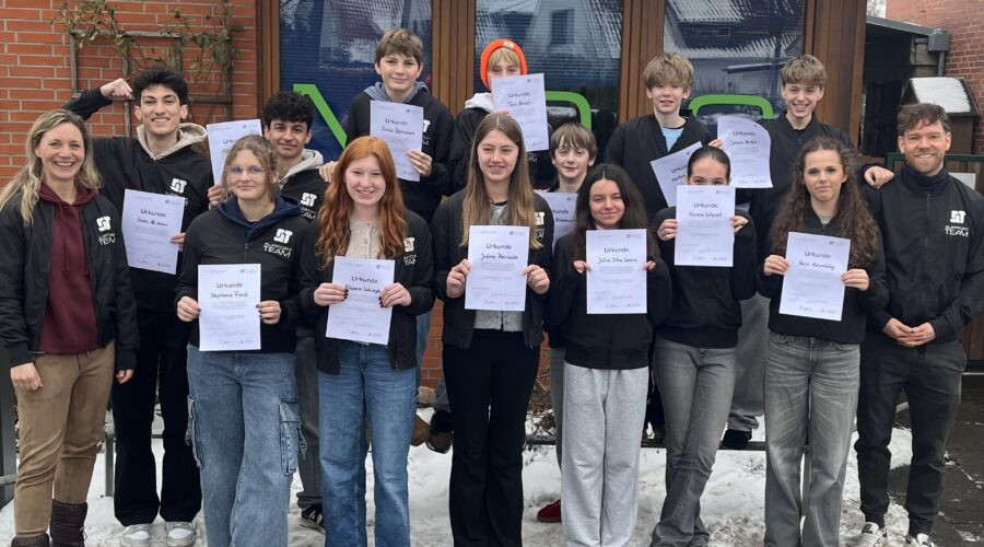 Gruppe von Jugendlichen hält Urkunden, lächelt vor einem Gebäude im Schnee.
