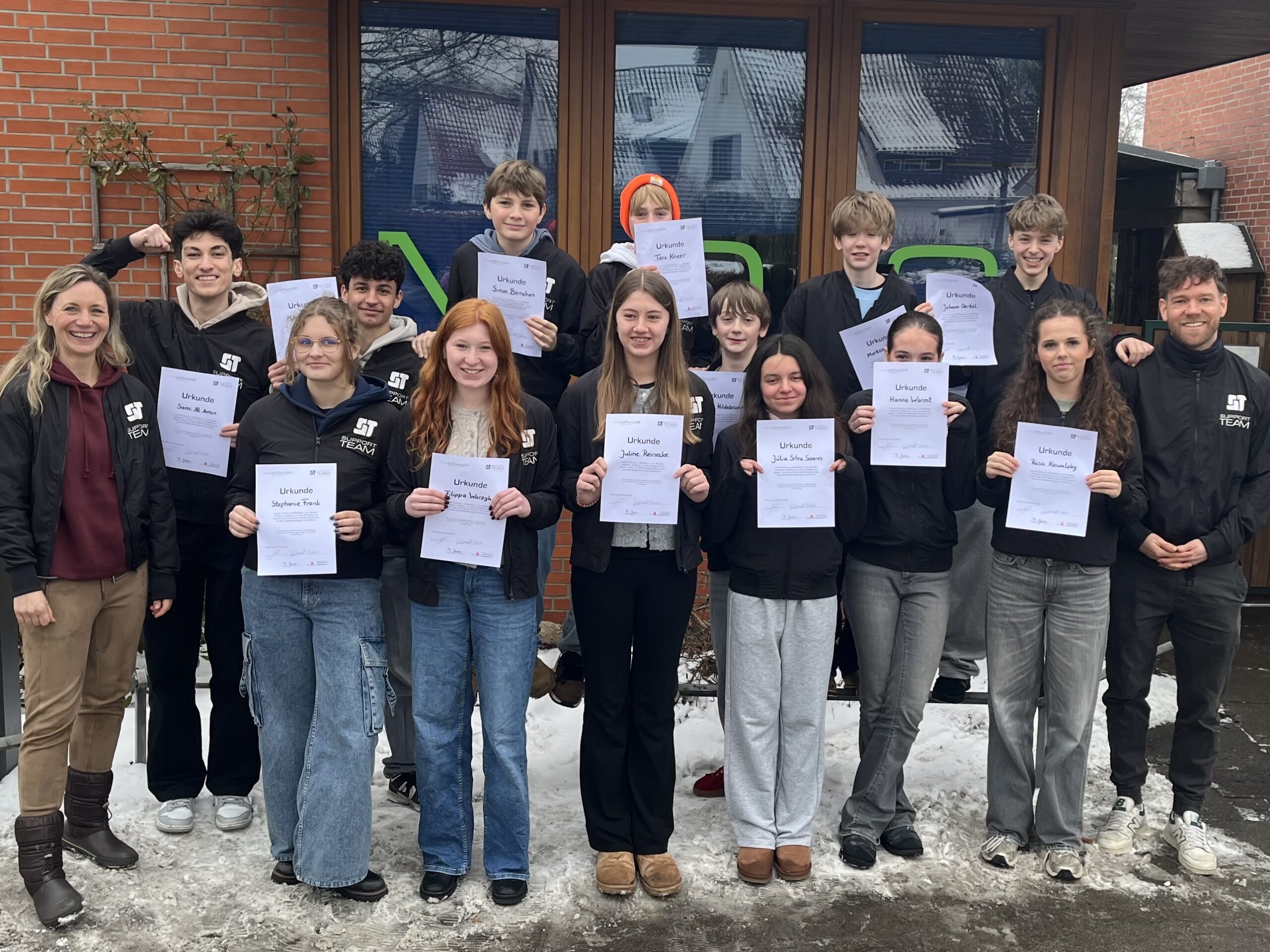 Gruppe von Jugendlichen hält Urkunden, lächelt vor einem Gebäude im Schnee.