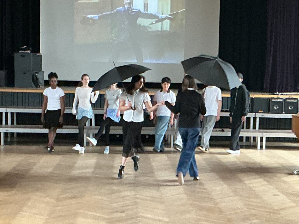Eine junge Frau mit einem Regenschirm tanzt auf einer Bühne, umgeben von anderen Personen.