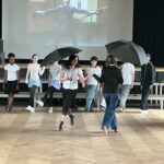 Eine junge Frau mit einem Regenschirm tanzt auf einer Bühne, umgeben von anderen Personen.